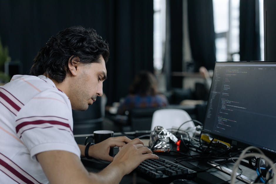 A young man with dark, curly hair is seated at a desk in a modern office environment, focusing intently on typing on a black keyboard connected to a large computer monitor displaying lines of programm